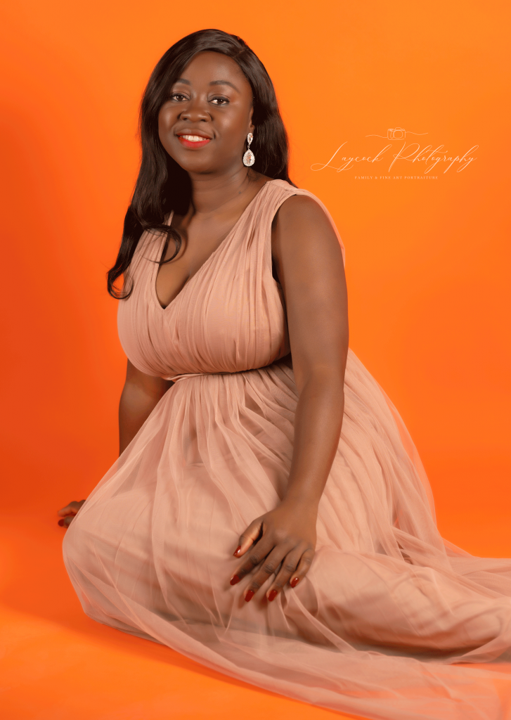African woman wearing a nude gown. Sat with legs out to the side on an orange background. Smiling, looking at the camera.
