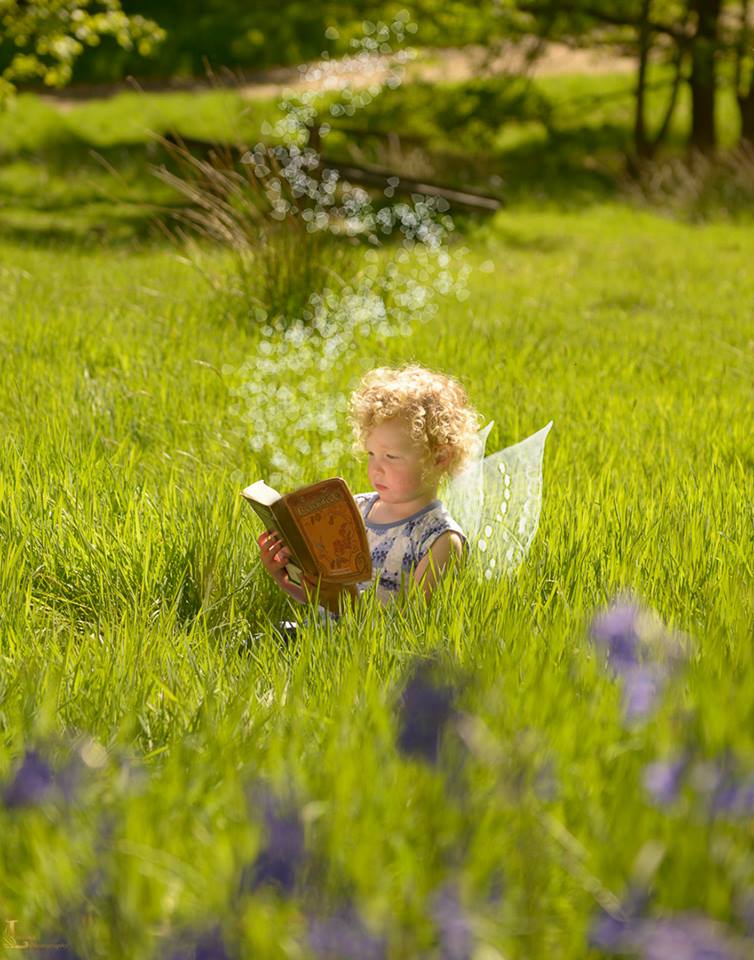 Small child sat i field reading a book. She has fairy wings on and bubbles coming out of the pages from the book.