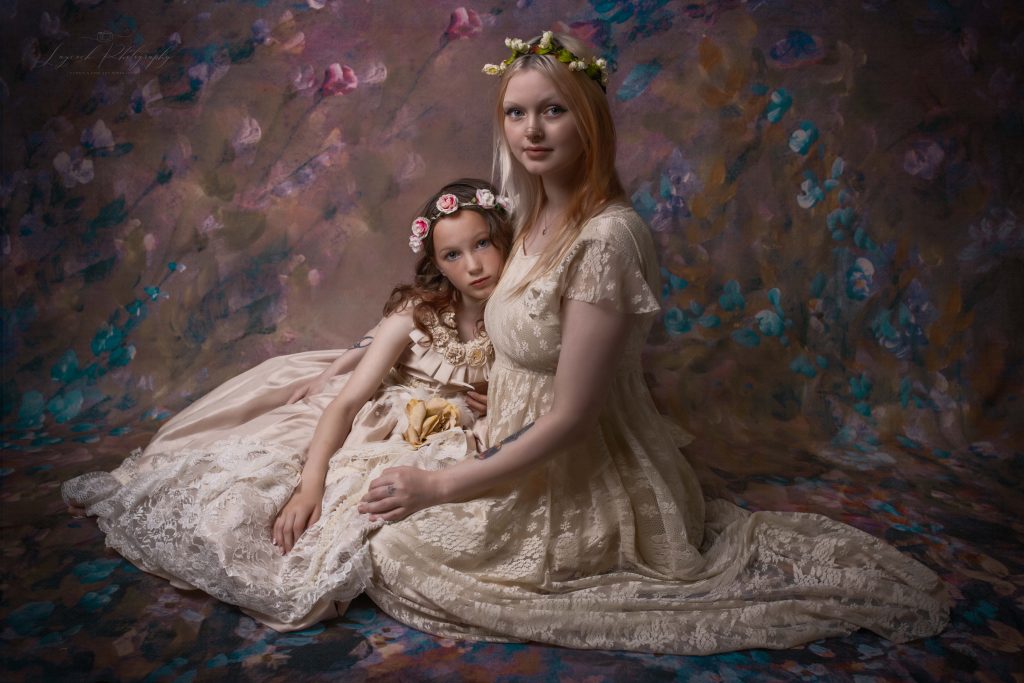 Mother and daughter fine art portrait in Stalybridge studio wearing elegant ivory lace dresses and floral crowns against a hand-painted floral backdrop.